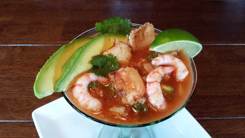 A photograph of a Chilean shrimp meat mexican cocktail with shrimp, avocado, lime juice, cilantro, tomato juice, clam juice, onions and cucumber
